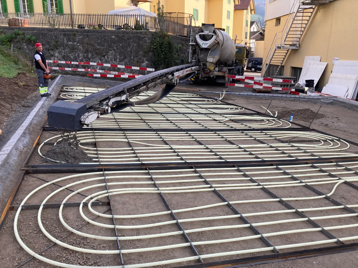 Strassenbauarbeiten. Man sieht weisse Kunststoffrohre, die kreisförmig angelegt sind, und einen Betonmischer, der sie mit einer grauen Masse bedeckt.